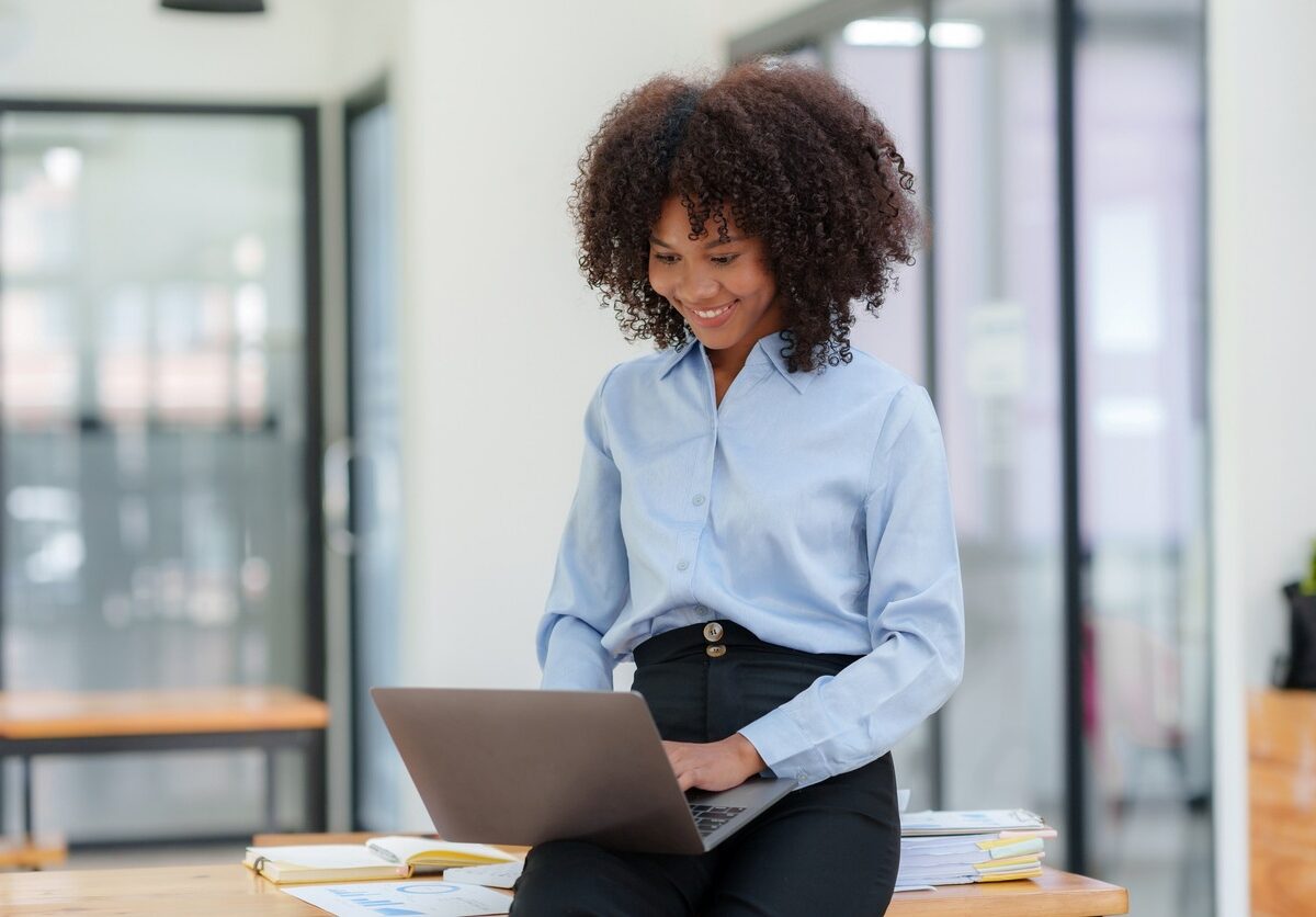 A woman in business attire smiling while working on a laptop in a modern office setting, seated on a desk.