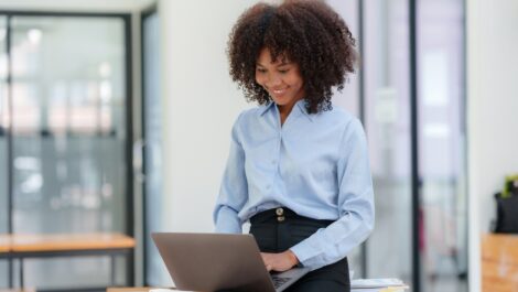 A woman in business attire smiling while working on a laptop in a modern office setting, seated on a desk.
