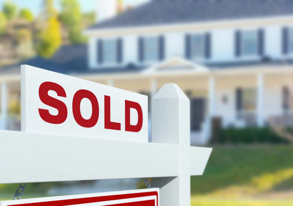 A red and white sign reading “Sold” sits outside the yard of a suburban blue and white home.