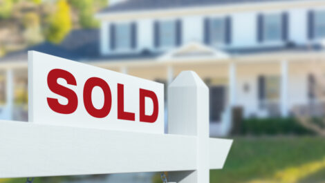 A red and white sign reading “Sold” sits outside the yard of a suburban blue and white home.