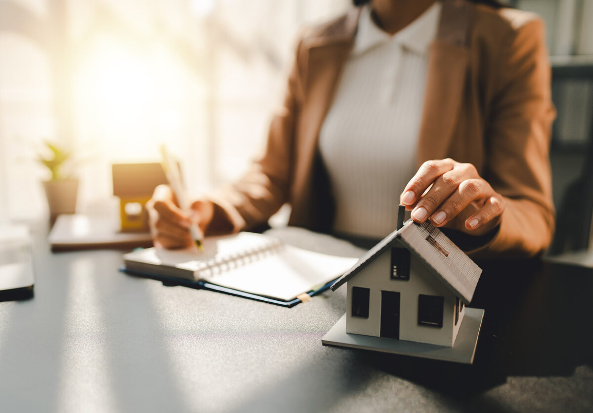 A real estate agent sitting at a desk, writing in a notebook, with a model house next to her.