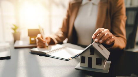 A real estate agent sitting at a desk, writing in a notebook, with a model house next to her.