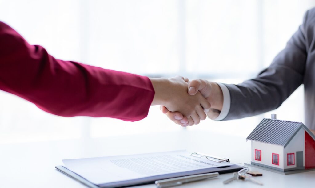 Two people shake hands after signing paperwork, with a model house on the desk next to the documents.