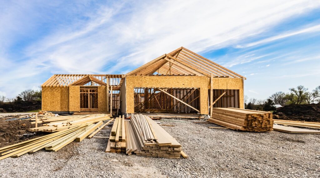 A home in the early stages of construction with long beams and floorboards assembled in the gravel on a sunny day.