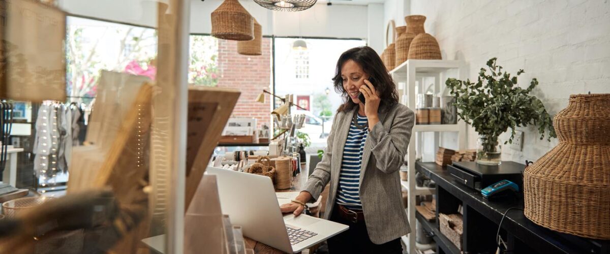 A woman stands behind the counter of a small business talking on the phone as she looks at a computer.
