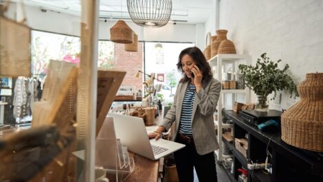 A woman stands behind the counter of a small business talking on the phone as she looks at a computer.